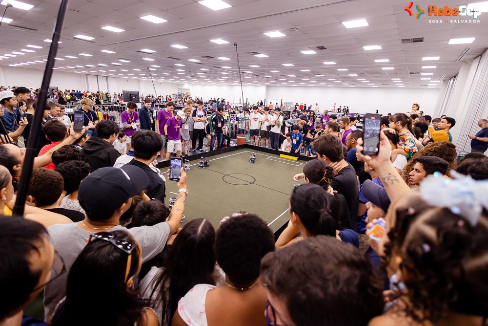 A large crowd, many holding up phones, surrounds a RoboCupJunior Soccer field during a match.