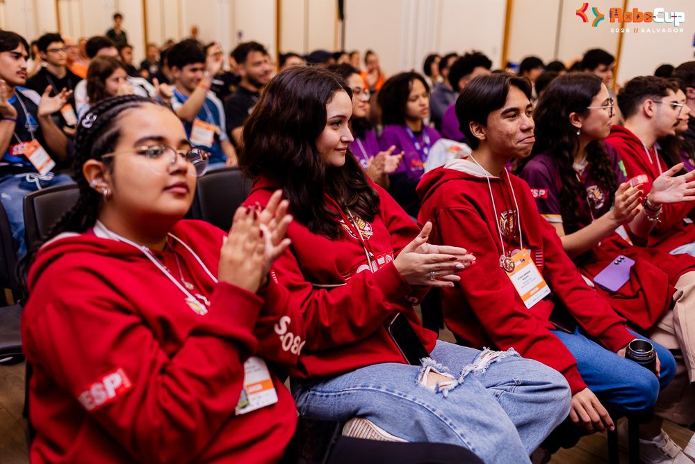 Students in matching red team hoodies clap and cheer in a packed auditorium.